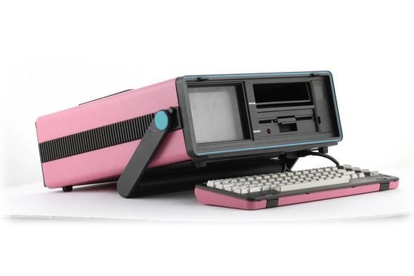 rosa pastel, amb una banda negra horitzontal amb ranures de ventilació a la part lateral. La pantalla, inclosa en un marc negre amb detalls blaus, és petita i rectangular, i mostra una superfície fosca —potser apagada o sense senyal.

A la dreta de la pantalla hi ha una unitat de disc flexible (floppy drive), també negra, amb una ranura oberta i un petit botó vermell que podria ser un indicador o interruptor. Un braç articulat de plàstic negre sosté la pantalla en angle ergonòmic.

El teclat, connectat per cable, té un carter rosa a joc amb el cos de l’ordinador i tecles grises de tipus mecànic, amb lletra negra. El disseny general és dels anys 80, amb formes arrodonides i colors vistosos, típics dels primers ordinadors domèstics o educatius.

L’estil és nostàlgic, tecnològic i visualment atractiu, ideal per a exposicions, col·leccions o continguts sobre història de la informàtica.