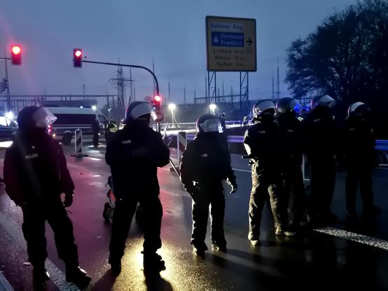 Eine Gruppe uniformierter, gepanzerter Polizeikräfte steht in Formation auf einer nassen Straße bei Dämmerlicht. Sie tragen Helme mit Visieren und wirken einsatzbereit. Hinter ihnen leuchten Verkehrsampeln rot, ein Polizeivan steht quer, und ein Wegweiser zeigt Richtungen wie Dortmund und Frankfurt. Die Szene ist von blauem Einsatzlicht und dunklem Himmel geprägt.