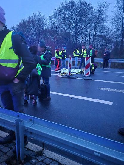 Mehrere Personen in gelben Warnwesten stehen auf einer nassen Fahrbahn am Rand einer Straße. In der Mitte liegt eine Person auf dem Boden, zugedeckt mit einer Rettungsdecke, während andere sich darum kümmern. Weitere Menschen beobachten die Situation, im Hintergrund Bäume im Dämmerlicht.