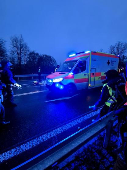 Ein Rettungswagen mit Blaulicht steht auf einer dunklen, nassen Straße. Mehrere Personen in Warnwesten und Einsatzkräfte befinden sich daneben. Im Hintergrund sind Bäume im Dämmerlicht zu sehen. Die Szene wirkt wie ein laufender Rettungseinsatz.