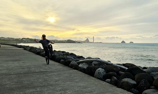Dude on a mountain-bike on New Plymouth's foreshore walkway giving a thumbs up while wheelying one handed past me. The sunset is glowing through the clouds behind, and high tide is breaking to the right. Paritutu Rock and the old power starting chimney at the port can be seen in the distance.