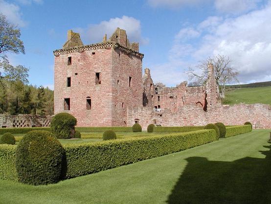 Edzell Castle, near Edzell in Angus. The image shows a pinkish stone ruined tower house across the centre of the frame, with the tower on the left and other buildings to the right of it. The tower is complete to roof level. This side of the buildings is a formal garden with low box hedges. The ground rises in the background. The scene is in sunlight.