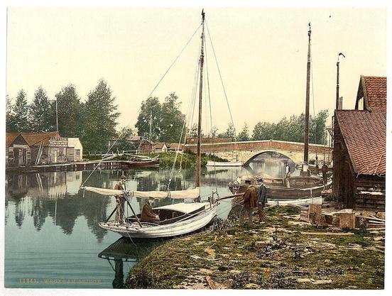 The image depicts a serene riverside scene with several boats moored along the bank. In the foreground, two individuals are seen on one of the small rowboats; they appear to be engaged in conversation or examining something together. A larger sailboat is visible near them, its sails furled and resting against the hull.

In the background, a stone bridge spans across the river connecting both banks. The surrounding area features traditional wooden houses with sloped roofs and various flags flying above some of these buildings. There are also tall masts rising from several structures along the waterfront, suggesting that this may be an active dock or marina. The sky is overcast, casting a soft light on the scene without creating harsh shadows.

The overall atmosphere exudes a sense of tranquility with people leisurely engaging in boating activities amidst picturesque surroundings indicative of late 19th-century England.