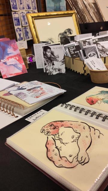 Photographie du stand, au premier plan un classeur à spirale avec des cartes postales petit monstre dedans, des fanzines au second plan, un petit pot avec des cartes de visites, un cadre avec un print