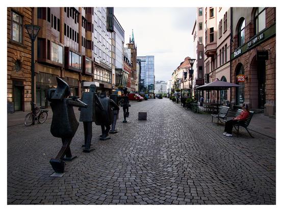 En liggande bild på Södergatan i Malmö med Optimistorkestern till vänster. Till höger på en bänk sitter en med hörlurar på huvudet. Höga hus med butiker på båda sidor denna gågata av kullersten.