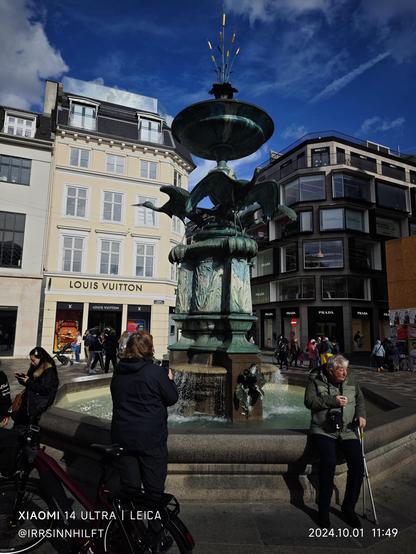 Die Sonne scheint am Storchenbrunnen in der Kopenhagener Einkaufsmeile.
Menschen flanieren und sitzen am Brunnen. 
Der Brunnen hat ein achteckiges Becken in dem ein Säulenaufbau steht,. Von diesem Aufbau "fliegen" Störche in alle 4 Himmelsrichtungen davon. Den oberen Abschluss bildet eine Schale mit stilisiertem Schilf.