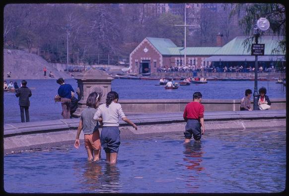 A group of people wading through water near a body of calm lake surrounded by buildings and trees. The individuals are standing in shallow waters, with some onlookers seated or walking along the shore. There is boat activity taking place in the background across the lake. Various boats can be seen, including rowboats and paddle boats, occupied by several people each. One individual appears to be piloting a small sailboat near the center of the image, but it's not under full sail at this moment. The architecture includes what looks like maintenance sheds or boathouses with signage on them. Trees in the background suggest that it might be late fall, winter, early spring, or possibly summer without leaves due to natural conditions such as climate change affecting foliage cycles.

The water has a serene and peaceful ambiance, reflecting some of the trees and structures around it. The color tone is slightly reddish-pink, indicative of older film stock used in capturing this photograph taken on April 1962 at Central Park, New York City.