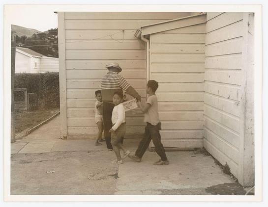 The photograph depicts a group of four individuals standing outside what appears to be the entrance to a house or storage shed. On the left, three children are visible; two boys and one girl who seem to be in mid-stride as if walking into or out of the building, while another child is slightly behind them. The child at the forefront holds up a sign with text on it but due to resolution limitations specific details cannot be made out.

On the right side stands an adult male wearing checkered shirt and dark trousers; he's holding onto one of the children who is carrying the aforementioned sign. He wears spectacles, a cap turned backward, and appears slightly stooped in his stance indicating perhaps some effort or weight being held up by him. His expression seems attentive towards something out of frame.

The backdrop includes a residential setting with a wooden structure that could be part of an extension to the house or storage facility they're entering/exiting from. The photograph is monochromatic, possibly vintage and has soft focus which suggests it may not have been taken on professional-grade equipment. No discernible brand markings are visible in this scene.

There's a subtle detail showing what looks like grassy terrain with more buildings faintly noticeable at the far end behind the structure where these individuals stand. The overall ambiance of the photograph feels candid, possibly capturing an everyday moment f [...]