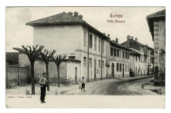 The image shows a black and white photograph of an old street scene, possibly from the early 20th century. It features European-style architecture with buildings that have pitched roofs and decorative details typical of Italian or Southern French towns. The streetscape includes narrow roads lined with variously sized windows, some adorned with shutters.

Two individuals are present in the image: one standing on the left side near a tree stump wearing suspenders, shorts, and shoes indicative of late 19th to early 20th-century fashion for men; another person is walking away from the camera towards the center-right part of the street. The setting appears tranquil with no modern vehicles or contemporary elements.

The photograph has text indicating "Turbigo - Viale Novara," suggesting it's a postcard depicting Viale Novara in Turbigo, which may be located within Italy given its architectural style and language used for writing on the image. A signature at the bottom left corner reads "Gasoll-Turbigo 19068." The overall condition of the photograph appears aged with creases and discoloration typical of old postcards from that era.

The reference provided indicates another related image, which is likely a larger or more detailed view of one element within this scene.