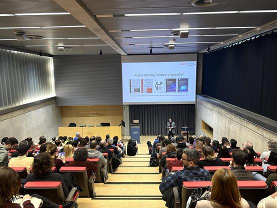 Olga Carreras a l'auditori de l'a11yConf 2025 a Girona parlant de european accessibility act davant de la audiència i assistents, amb un pla més ampli amb la sala plena d'assistents