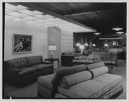 The image depicts an interior space, likely a showroom or display room for furniture. The scene is composed of various pieces of upholstered seating arranged in different configurations: couches and love seats are the most prominent items visible. They feature ornate patterns with plush fabric upholstery.
A large framed painting hangs on one wall above a sofa; it appears to be historical, featuring figures from an earlier era dressed in what could be 18th-century attire. The room is well-lit by multiple lamp fixtures overhead and standing beside the furniture pieces. Rugs are spread across different areas of the floor, adding warmth and texture.
The flooring itself seems consistent throughout; it's a patterned carpet with geometric designs that harmonize with the style presented in this setting. In terms of color tones, due to its black-and-white nature, the photo predominantly features shades from greys to blacks along with some muted earthy hues on the furniture upholstery and rug patterns.
Overall, there is no visible activity or people present within the scene; it focuses solely on showcasing the furnished items in an orderly layout. The absence of modern digital devices indicates a vintage setting likely dating back several decades ago based upon style cues such as lamp design and overall room aesthetics.
