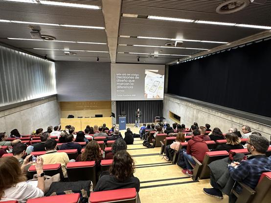 Berta Nicolau a l'escenari de l'auditori de l'a11yConf 2025 a Girona parlant de disseny i accessibilitat digital davant de la audiència, amb un pla més ampli amb la sala plena d'assistents
