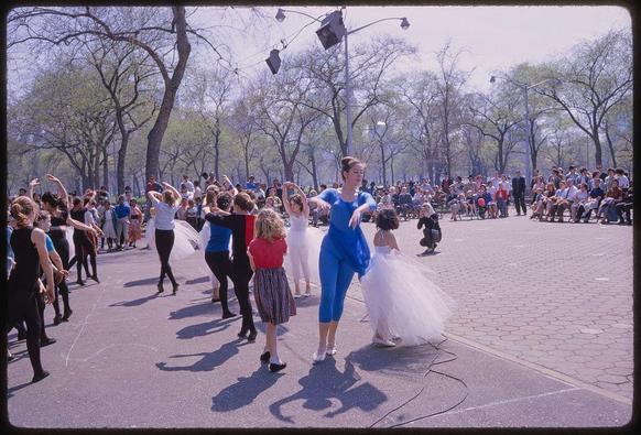 Image showing a lively scene in what appears to be a public park. In the foreground is Toni Frissell, dressed in blue with her hair pulled back, mid-twirl as she dances amidst various people who are also dancing around her. They seem engrossed and joyful. Most of them wear casual clothes; some women have on tutus or skirts while others don pantsuits. The setting has a paved area surrounded by trees without leaves suggesting it could be springtime in the northern hemisphere. In the background, there's an audience seated along lampposts watching this dance event unfold with apparent interest and amusement.