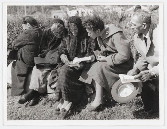 The photograph is a black and white, grainy image that captures a candid moment among five individuals seated on the grass. Four of them appear to be women while one seems to be a man based on their clothing style which includes suits with ties, coats, and hats— indicative of an earlier time period possibly mid-20th century attire.

Three of these women are dressed in winter or fall outerwear; two have fur collars over heavy woolen dresses. Their attention is focused outwardly as they seem engaged in conversation rather than looking at the camera which suggests a casual, unposed moment captured perhaps during an event outdoors on what appears to be a bright sunny day given their shadows and clothing choice for warmth.

The man seated between two women has his arm resting casually across the back of one woman's chair. He is holding papers or possibly tickets in his hand while another document rests atop his right knee suggesting they might have been attending an event, perhaps a fair where pamphlets were distributed as seen from their hands and documents spread out before them.

The background shows lush greenery which could indicate that the photo was taken at a park during spring or summer. There is no definitive indication of age range but it seems to be mature adults possibly in late middle ages judging by wrinkles visible on faces, particularly prominent around eyes for female subjects.

The image carries an air of h [...]