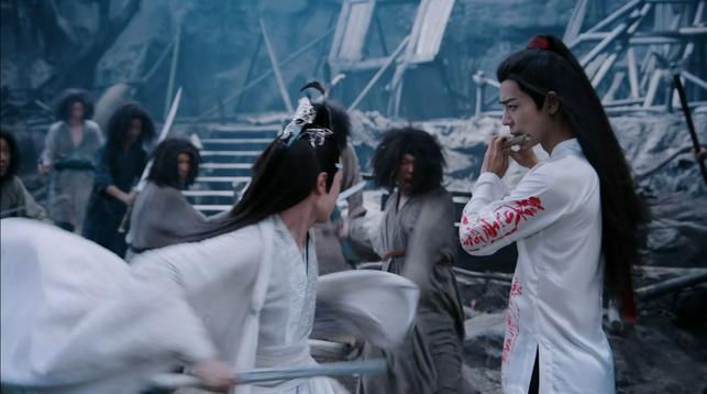 Wei Wuxian (actor Xiao Zhan) (in white shirt (with red writing) playing a dizi while Lan Wangji (actor Wang Yibo) (in white robes) is fighting around him with a sword to keep the puppets away.