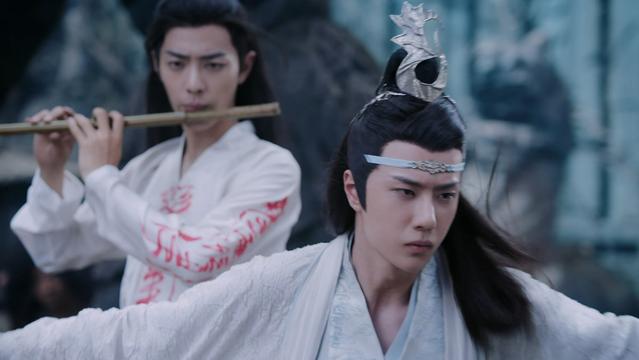 Closrup of Wei Wuxian (actor Xiao Zhan) (in white shirt (with red writing) playing a dizi while Lan Wangji (actor Wang Yibo) (in white robes) is fighting around him with a sword to keep the puppets away.
