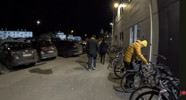 A person wearing a yellow toque and a puffy yellow jacket is unlocking their upright bike, which is parked with other upright bikes at a wheelbender rack on Bodø, Norway. People walk by in the night, all leaving the stadium.