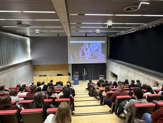 Mia Salazar en l'escenari de l'auditori de l'a11yConf 2025 a Girona parlant d'atributs ARIA davant de la audiència, amb un pla més ampli amb la sala plena d'assistents