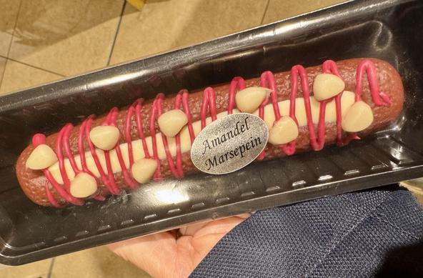 A dessert in a plastic container featuring a long, almond-flavored marzipan treat decorated with pink icing and almond pieces on top. A label reads "Amandel Marsepein."

frikanel aus marzipan