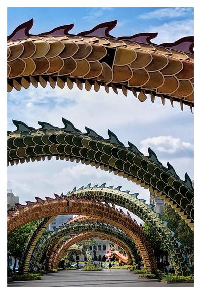 Photo of a series of swooping arches over a wide pedestrian avenue receding to a building in the distance. The arches have scales and fins in hues of orange and green, and the arch at the far end bears a sculpted dragon’s head. The effect is of a dragon’s long body encircling the avenue, leading the eye to a statue of a man with one arm raised (Vietnamese leader Ho Chi Minh) in front of a palatial facade. Trees and shrubs line the sides of the avenue, and the sky seen through the dragon arches is blue with white clouds.