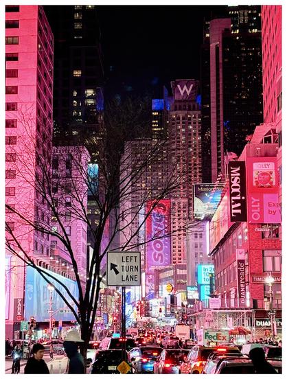 A nighttime view of Times Square, NYC, looking down 7th Avenue from around 50th Street. Traffic flows in a river of red taillights through a canyon of buildings awash in pink light. In the foreground stand a leafless tree and a sign that reads LEFT TURN LANE with a helpful arrow. Pedestrians cross the street at bottom frame and mill about the sidewalks. Signs and banners project from the sides of buildings, most prominently a pink vertical ad that reads SONIC3 (for the “Sonic the Hedgehog” sequel). In the distance is the high-rise building of the Marriott W Hotel, with a large “W” sign at the top.