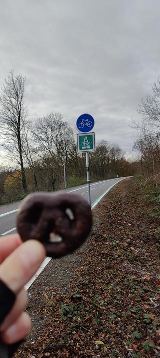 Meine Hand hält eine Lebkuchenbrezel ins Bild. Dahinter ist ein Radschnellwegschild vor selbigem zu sehen