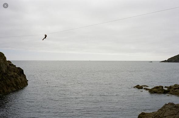 Maria hängt weit oben an einer Zipline über einer felsigen Meeresbucht; grauer Himmel, ruhiges Wasser, Felsen links und rechts im Vordergrund.