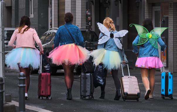 Four young women in neon-colored tutus with jeans jackets in blue, aqua and pink, pulling trolley cases and walking away from the camera.
