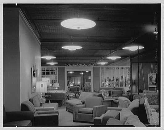 The image depicts a spacious interior, likely of a commercial establishment such as a furniture store or showroom. The room is well-lit with multiple round overhead lights casting an even glow across the space. Several pieces of upholstered seating are arranged throughout, including sofas and armchairs in various shades and styles. Some chairs have throw pillows on them.

On one side of the room stands tall display cases filled with what appears to be books or magazines, providing a sense of depth to the scene. The walls feature framed artwork or photographs that contribute to an aesthetically pleasing atmosphere. A large window is visible in the background, offering natural light and possibly connecting this interior space with other parts of the building.

The image seems vintage, as indicated by its monochrome tone and style, suggesting it was taken sometime around 1950s May. It appears to be a photograph from Ludwig Baumann's business located on Newark Street in New Jersey, specifically found on the fourth floor during an unspecified date mentioned earlier that year (May). This information is attributed to Gottschoschleisner Inc., which might have been involved with photography or archiving at that time.