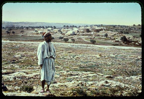 The image depicts a man standing on a rocky, arid landscape. He is wearing traditional Middle Eastern clothing: a long white garment with dark vertical stripes around the waist and cuffs, paired with sandals that appear to be of a simple design. The attire suggests he may come from an agrarian or pastoral background. On his head, he wears what seems like a turban-like cap. In one hand, he holds a stick which could serve as a walking aid or perhaps for managing animals.

Behind him is an expansive view of rural settlement with numerous structures, possibly huts and homes. The architecture indicates that this may be in the Middle East or North Africa region. Sparse vegetation dots the landscape; notably, there are some trees scattered around but no lush greenery indicative of a fertile farmland. In the distance, hills can be seen under a clear sky.

The photo has an aged look to it, with slight discoloration and fading, which is characteristic of older photographic prints. This could suggest that the photograph was taken many years ago when color film technology might not have been as advanced or prevalent for such images. The overall ambiance conveyed by this image evokes a sense of solitude amidst rural life in what appears to be an ancient settlement area.

The caption suggests additional historical context, indicating the photo is from "Northward from Jerusalem" and it could possibly link to biblical narratives given i [...]