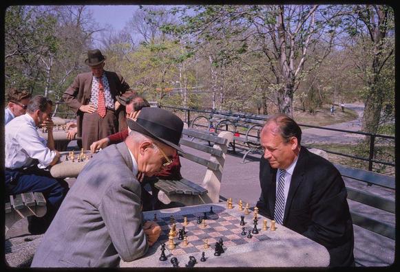 A group of men playing chess in a park, with trees visible in the background and other people seated at tables behind them. The image appears to be taken from Central Park during April 1962 by Toni Frissell. This photograph is part of her photography collection that includes landscapes and cityscapes captured throughout New York City, particularly focusing on Central Park.