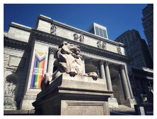 Photo of the exterior of the main research branch of the New York Public Library. The sunlit statue of Patience the lion sits on its stone pedestal in front of the library’s grand Beaux-Arts marble facade, with its columns, arches, and frieze of sculpted figures. On the left and right sides of the building are tall vertical banners bearing the rainbow design of the Pride flag, over which are printed the words LIBRARIES ARE FOR EVERYONE and PROTECT THE FREEDOM TO READ.