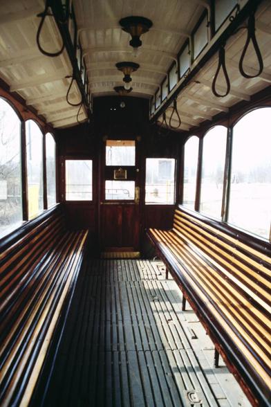 Foto im Hochformat. Die Kamera blickt in einen Straßenbahnwagen mit Holzbänken, die seitlich unter den Fenstern entlang laufen. Oben hängen Halteschlaufen.