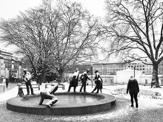 Eine verschneite Szene mit einer Brunnenanlage, in der mehrere lebensgroße Bronzefiguren posieren. Im Hintergrund stehen kahle Winterbäume und städtische Gebäude, während eine Person in warmer Kleidung am Becken vorbeigeht. Alles liegt unter einer dünnen Schneeschicht und wirkt ruhig und klar im Winterlicht.