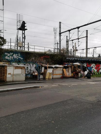 A row of well built slum houses made of recycled material, mostly wood, along a train track.
To the right, a man is working on the extension of his dwelling.