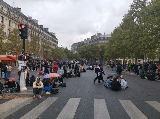 A wide street in Paris. No vehicles. People sitting on the ground in small groups.