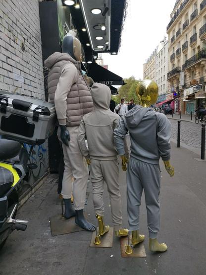 Three golden shop mannequins : one male and two children, on a sidewalk, viewed from behind.