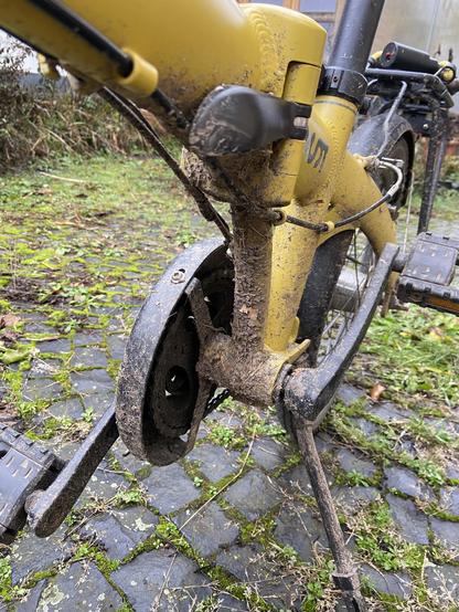 Blick unter ein gelbes Faltrad. Der Rahmen rund um das Tretlager ist von einer dicken Dreckkruste überzogen. Das Rad steht im Freien auf Kopfsteinpflaster zwischen dem Moos wächst.