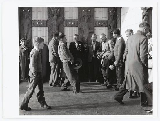 The black and white photograph depicts a group of people gathered indoors, with intricate wooden carvings adorning the walls in the background. The individuals are dressed in formal attire, suggesting an event or gathering possibly from mid-20th-century Europe based on their clothing style. Among them stands out a young boy casually walking towards the camera, contrasting his relaxed demeanor against others who appear engaged in conversation and activities such as holding what seems to be vinyl records. Notably absent is any indication of modern technology like smartphones or contemporary fashion trends, reinforcing its historical context. The image captures candid expressions of curiosity, engagement, and casual interaction among a diverse group of individuals.