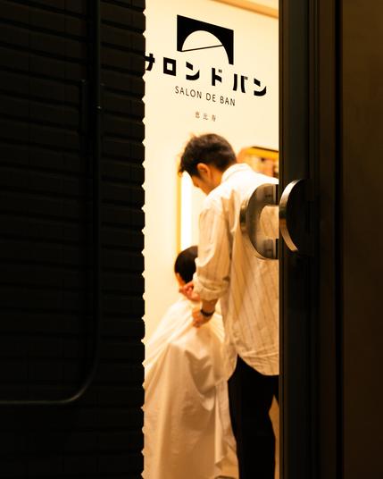 A barbershop interior seen through a partially open door, with a person in a striped shirt attending to a seated customer covered by a white salon cape. "Salon de Ban" is written on the wall, along with stylized Japanese text and a logo featuring a semicircle. Dim, warm lighting gives a cozy ambiance.