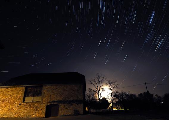 Trainées d'étoiles (poses de 4 s. pendant 20 minutes) avec au premier plan une vieille grange sous éclairage urbain et une lune qui dépasse l'horizon entre deux arbres sans feuilles.