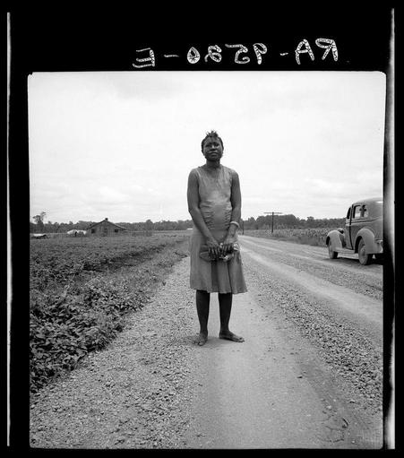 The image is a black and white photograph depicting an individual standing on what appears to be a dirt road. The person, who seems to be wearing a sleeveless dress or apron over shorts, has bare feet and holds a pair of shoes in their hands.

Behind the individual, there's a rural scene with fields that have been tilled or harvested, visible crops growing alongside some patches of soil without vegetation. A vintage car is parked on the side of the road, indicating an earlier time period when such vehicles were common.

Above the photo, handwritten text reads "3-0627-A9," which could be a date and identifier for this specific photograph within a collection or archive. Additional marks resembling symbols appear at different positions above the image as well.

The overall setting suggests agricultural labor in a rural area during what appears to have been an earlier era of American history, possibly relating to historical documentation on farming life, migrant workers, or similar social themes documented by photographers like Dorothea Lange.

(Note: The specific context and background story about this photograph are not provided within the image itself. Any additional information would require external research beyond what is visible in the photo.)