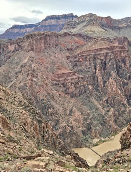 A view of a rock wall within the Grand Canyon where newer layers of horizontal sedimentary rock lie atop older, more vertically oriented metamorphic rock. A bridge across the Colorado River is visible at the bottom.
