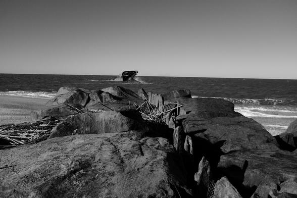 Monochrome image of the rectangular profile of a shipwreck a few hundred feet offshore. The wreck is a portion of the broken hull, and lies on its right side in the surf, facing away from the viewer.