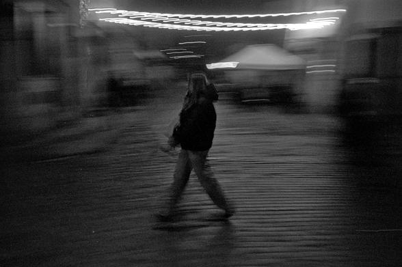 The image shows a blurred nighttime street scene captured using ICM (Intentional Camera Movement). A lone figure walks across a cobblestone surface, their form stretched and softened by motion, giving them a ghostlike, transient presence. Overhead, strings of lights streak into fluid, wavering lines, adding a sense of rhythm and energy to the composition. The surrounding buildings dissolve into dark, indistinct shapes, emphasizing the feeling of movement and fleeting passage. Overall, the photograph conveys a dreamy, cinematic atmosphere — a moment suspended between motion and stillness.