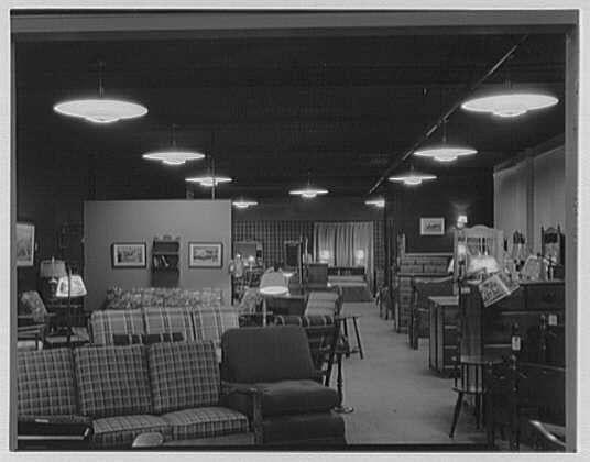 The image is a black and white photograph depicting an interior space, likely from the early to mid-20th century. The setting appears to be a furniture showroom or store with various pieces of upholstered seating arranged throughout the room in different patterns such as checks and stripes. There are multiple hanging lamps casting light on the displayed items.

On one side, there is shelving housing books, boxes, and possibly other merchandise, while another section shows what looks like artwork hung on walls behind display cases or counters. The overall ambiance of the space suggests a commercial setting with an emphasis on providing customers various options for furnishing their homes.

The photograph seems to be taken from one end of the showroom looking down its length towards where it presumably ends and continues beyond the camera's view, creating a perspective that draws attention into depth.