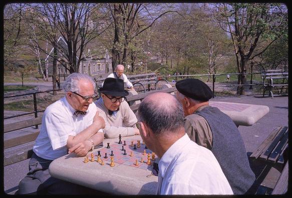 A group of four elderly men play chess in a park. They are seated around a portable table with an outdoor checkerboard setup, and the players use traditional black and white pieces on alternating squares.

Two of the men wear glasses; one has a bowtie under his shirt collar while another wears a sweater over a collared shirt. Their attire is casual but neat, suggesting they value their appearance despite their age. The man with the bowtie gestures animatedly as he discusses his next move in the game.

In the background, there are other people seated on park benches and walking through an area of lush greenery and trees typical of a city park during spring or early summer months.

A small church-like building is visible among the foliage, contributing to the relaxed atmosphere. The sky above is clear blue with very few clouds, indicating pleasant weather conditions at the time the photo was taken.