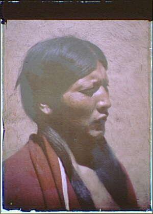 The image depicts a person with dark hair and features typical of indigenous American Indian characteristics, including facial contours like high cheekbones. The individual is wearing what appears to be traditional clothing or garments; specifically, they have on red outerwear that suggests it may be ceremonial attire. Behind the subject's head is an indistinct background which provides no contextual information about their environment.

The photograph carries a vintage quality indicative of early color photography during the late 19th and early 20th century in America. The name "American Indian" along with references to Acoma, New Mexico between 1906-1928 suggests this image may have been taken among Native American tribes living there.

This photograph is part of a larger collection by Arnold Genthe known for his work that includes portraits and documentary photographs depicting various cultures in the United States during those periods. The particular mention "autochrome" refers to an early color photography method, adding more historical context about this image being one of relatively few examples from such technology's era.

The photograph is presented on a website which hosts collections of archival material including artworks like this photo and others related to Genthe’s oeuvre or other significant cultural subjects.