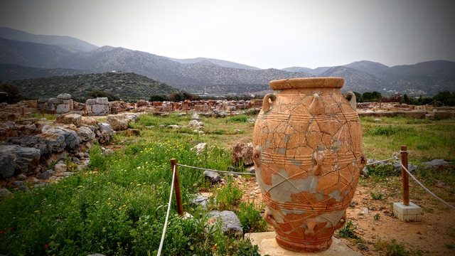 Auf einer Fläche, die teils mit Gras bewachsen ist, steht eine große Amphore von fast 2m Höhe. Auf der Fläche erkennt man Fundamente und Mauerrest eines Palastes und kleinen Stadt. Auch die Amphore scheint aus Scherben zusammengeklebt worden sein.
