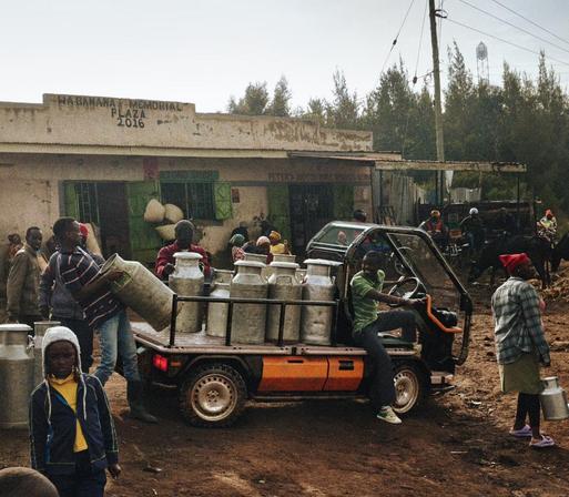 A bustling market scene with people engaging in various activities. A small vehicle loaded with milk cans is parked in the foreground, while several individuals, including children and adults, are seen around the vehicle and the nearby shop.